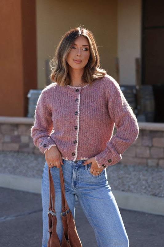 Woman in a Pink Speckled Button Down Cardigan and light blue jeans holds a brown bag outdoors.
