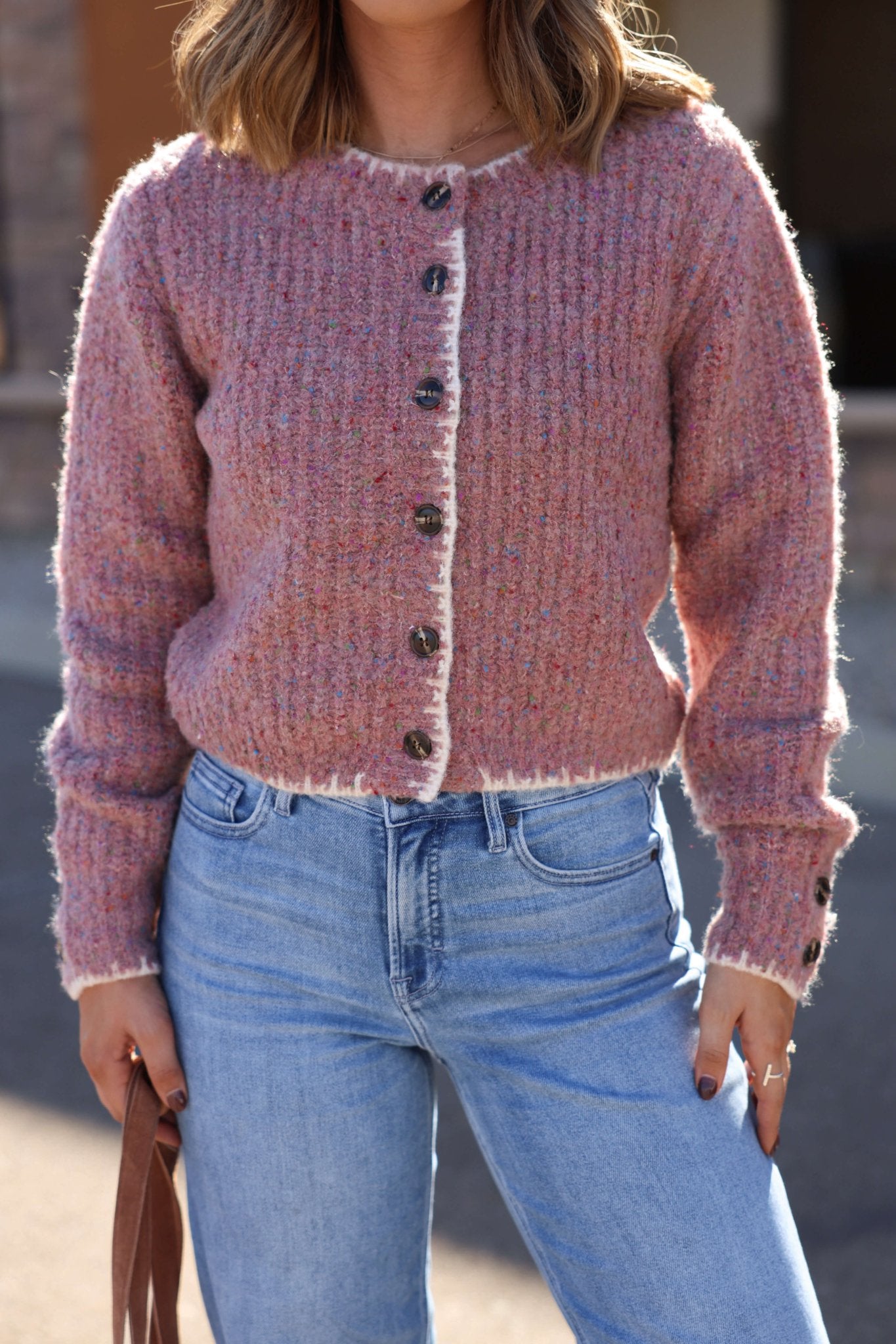 Woman in a Pink Speckled Button Down Cardigan and light blue jeans holds a brown bag outdoors.