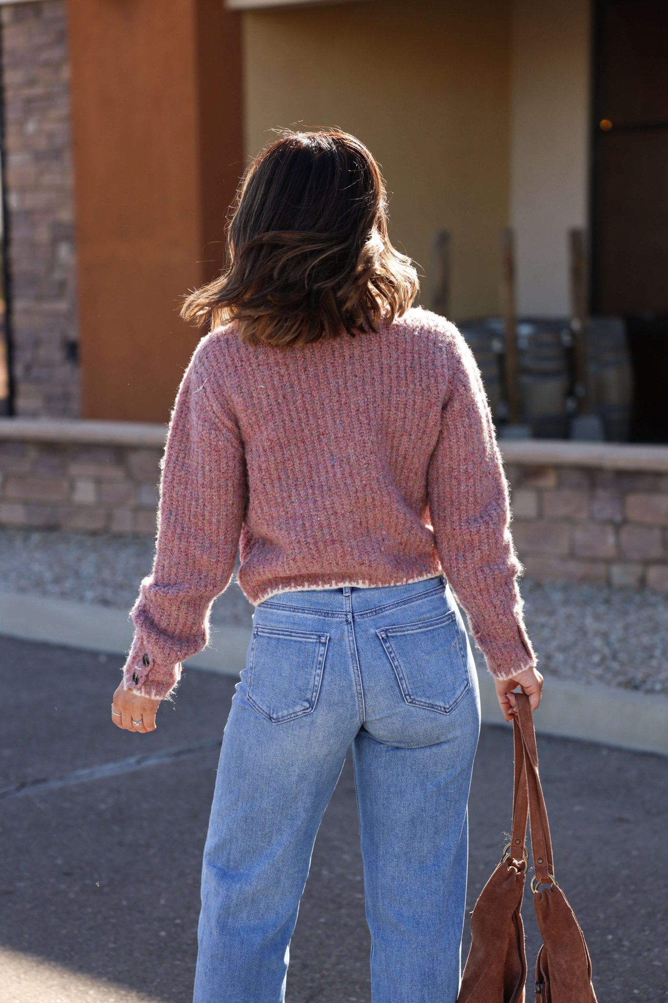 A woman in a Pink Speckled Button Down Cardigan and blue jeans stands outdoors with a brown bag, facing away.
