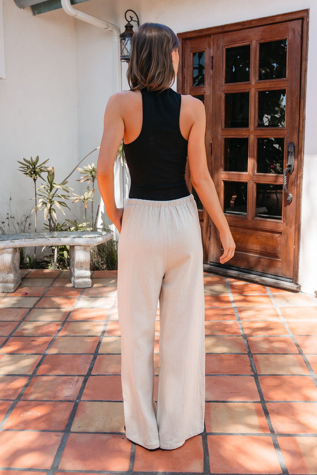 A woman stands outside on a tiled patio, wearing Piped Drawstring Linen Pants - Beige and a black sleeveless top.
