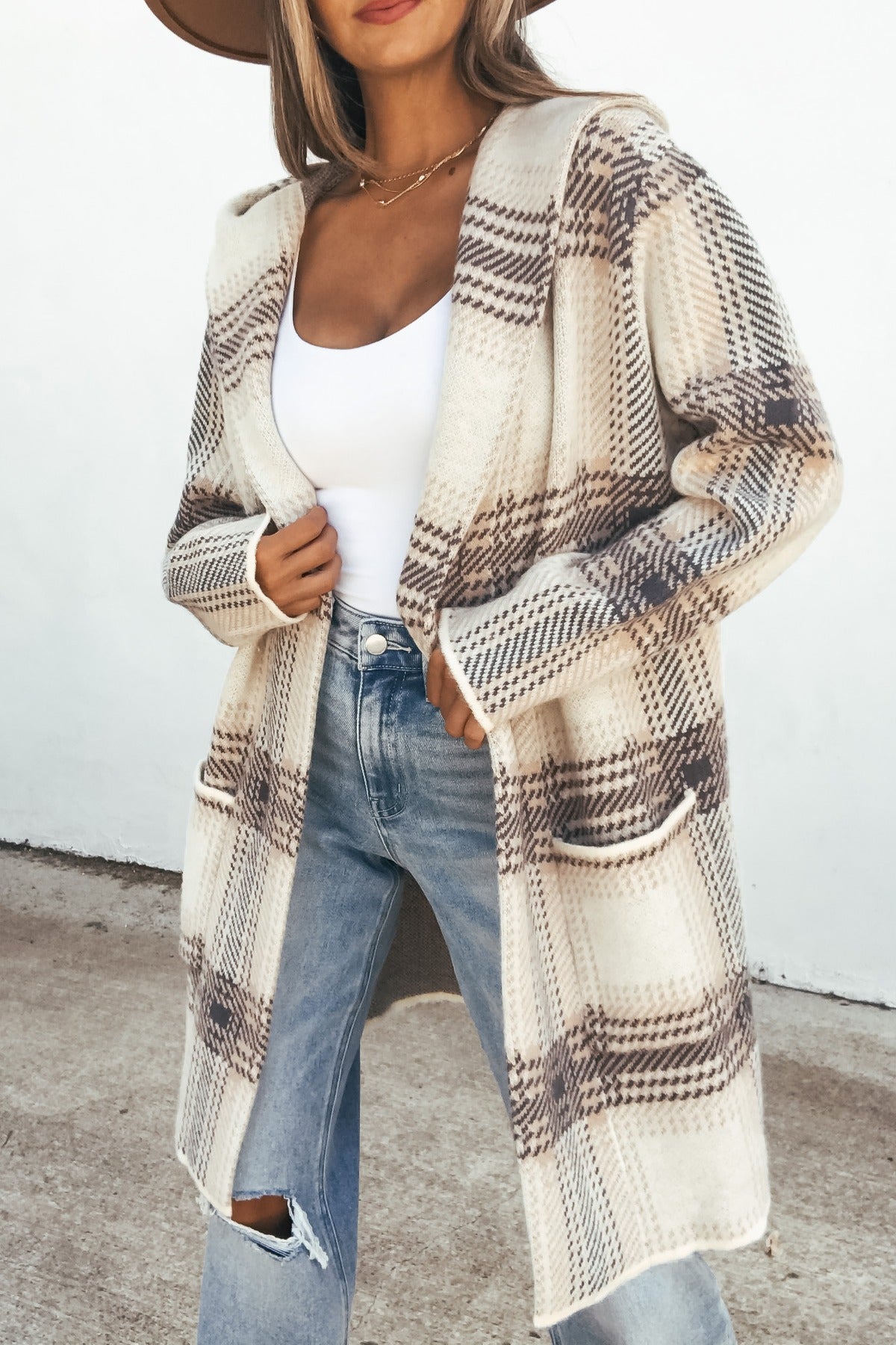 Woman in a Plaid Open Front Hooded Cardigan - Ivory with a white top, ripped jeans, and wide-brim hat against a light background.
