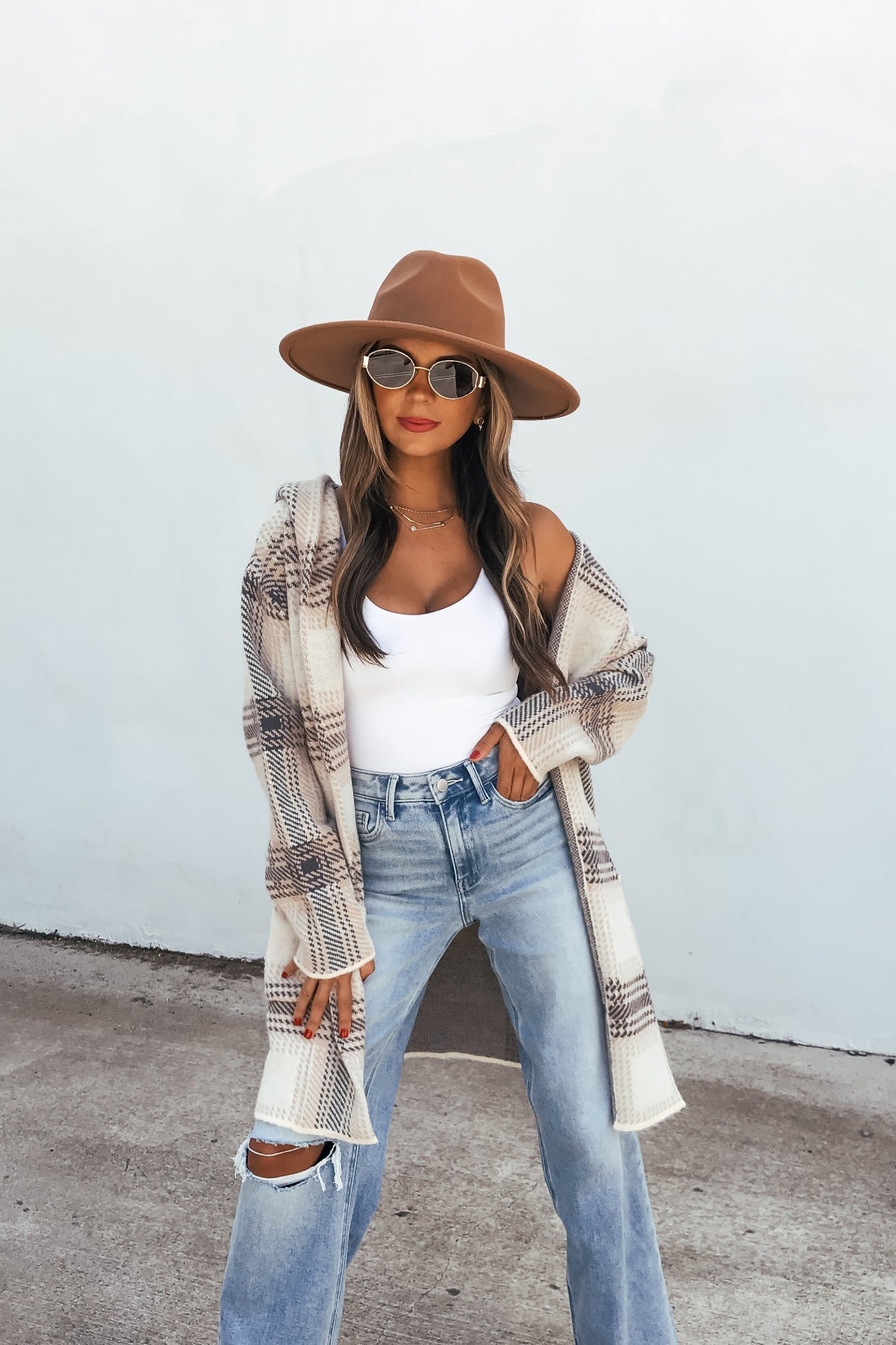 Woman in wide-brimmed hat, sunglasses, Plaid Open Front Hooded Cardigan - Ivory, white top, and ripped jeans stands by a light wall.