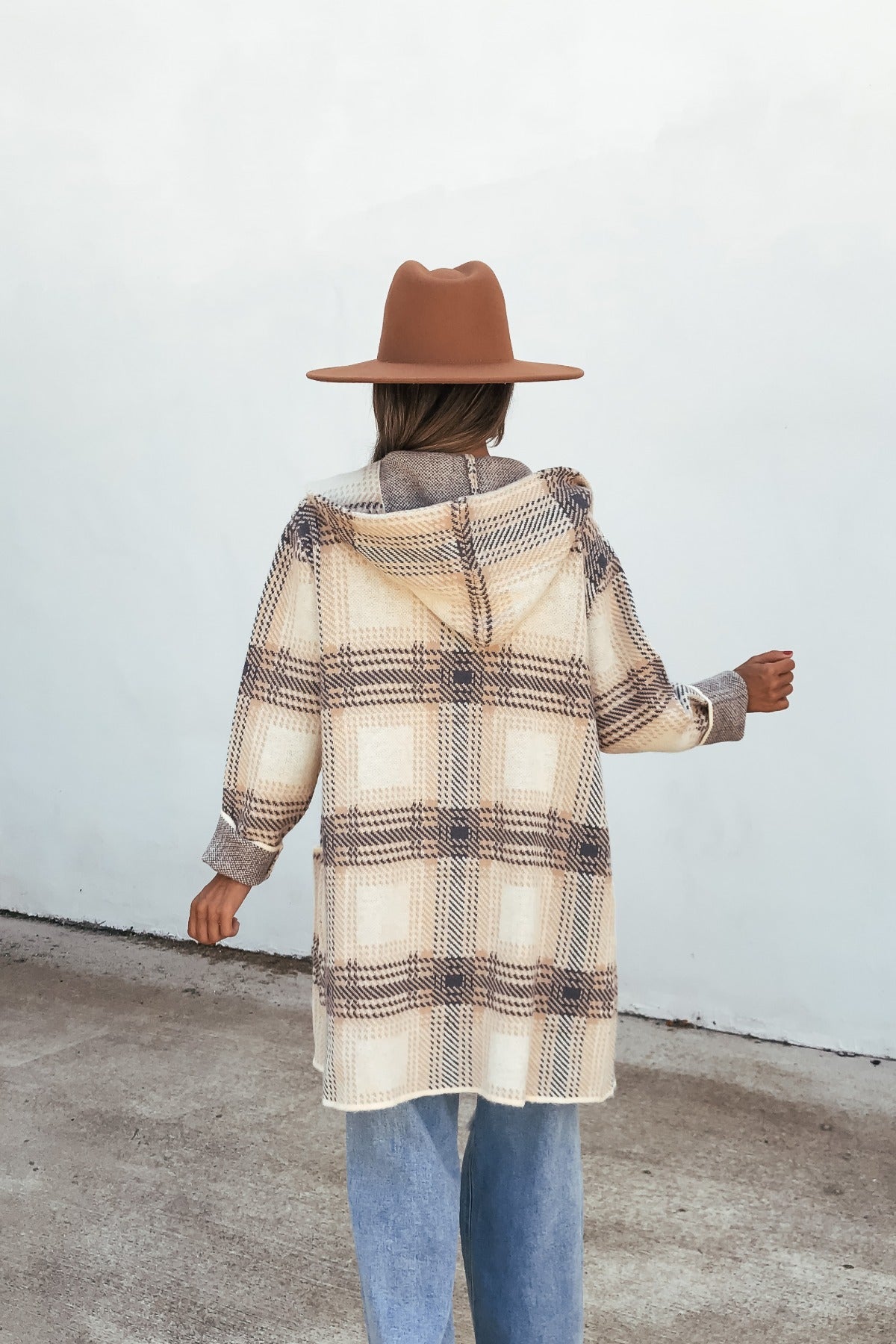 Person in a brown hat wears the Plaid Open Front Hooded Cardigan - Ivory, light blue jeans, and stands outdoors facing a white wall.