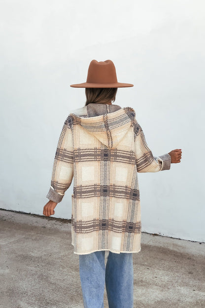 Person in a brown hat wears the Plaid Open Front Hooded Cardigan - Ivory, light blue jeans, and stands outdoors facing a white wall.