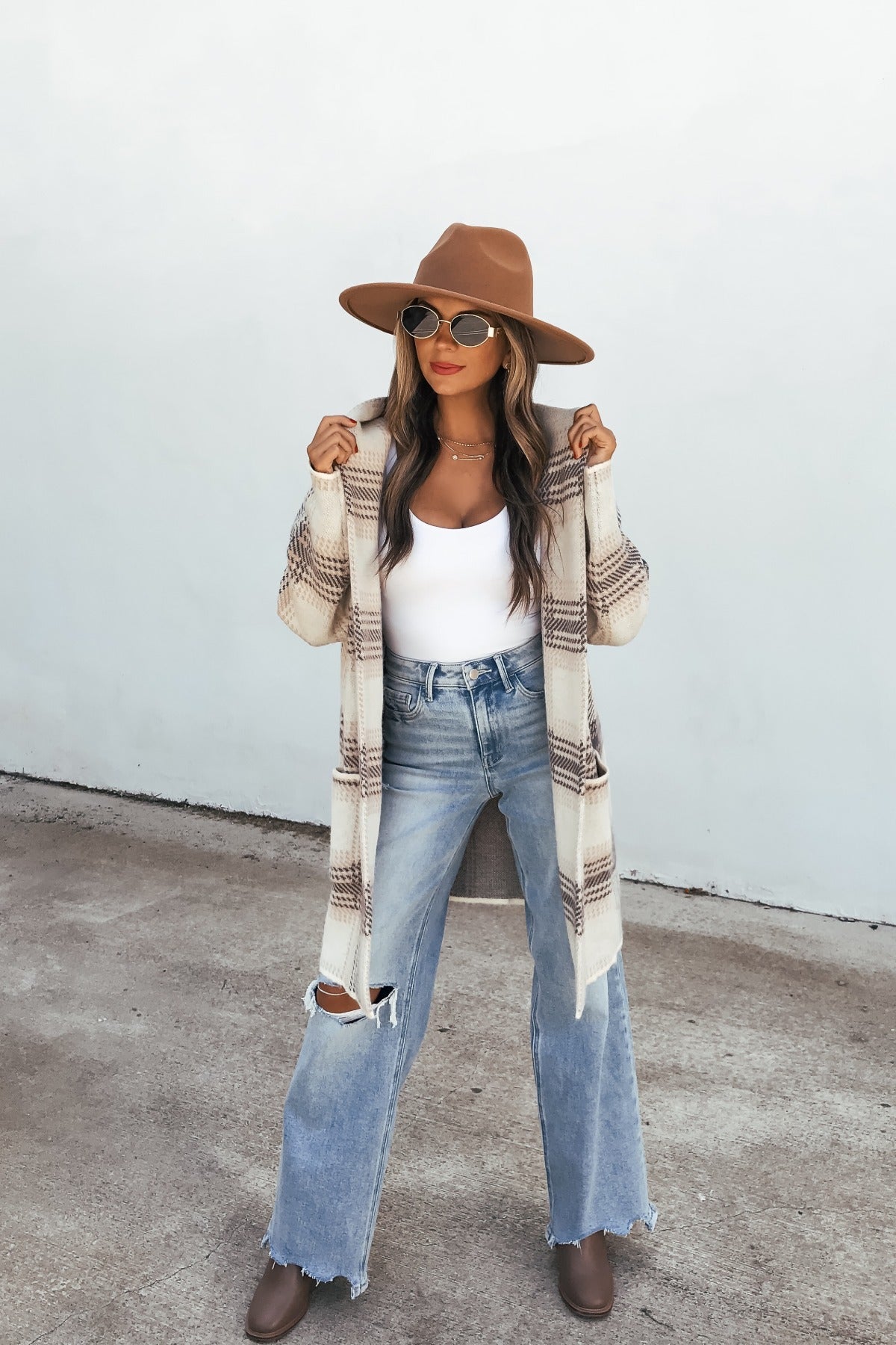 Woman in Plaid Open Front Hooded Cardigan - Ivory, brown hat, white top, ripped jeans, and boots stands by a plain light wall.
