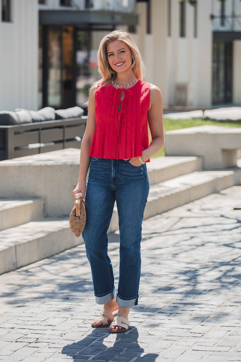 Woman outdoors in a Pleated Tie Front Top - Red, blue jeans, sandals, and a small woven bag, smiling at the camera.