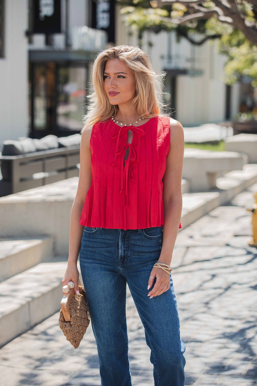 A woman in a Pleated Tie Front Top - Red and blue jeans stands outdoors, holding a woven clutch before modern buildings.