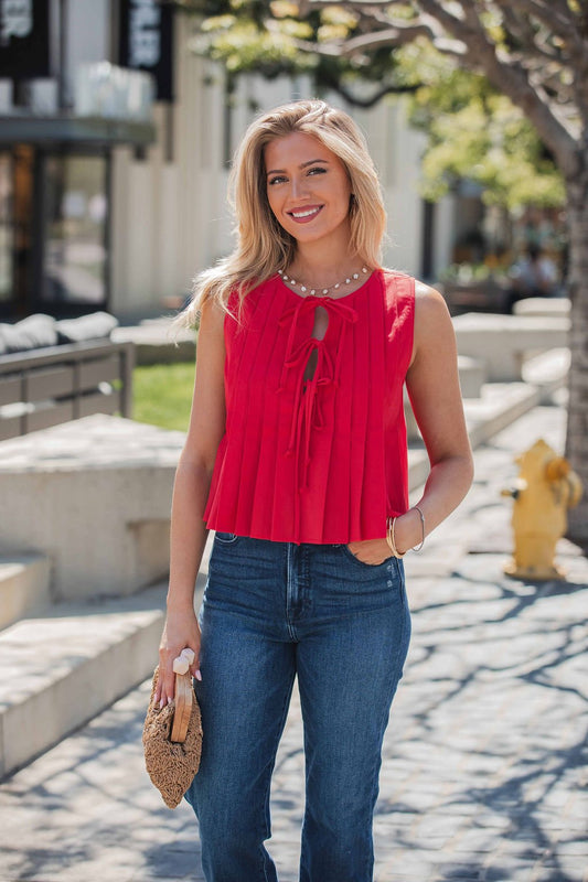 A woman in the Pleated Tie Front Top - Red and jeans stands outdoors, smiling with a woven clutch, trees and buildings behind her.