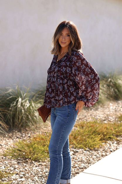 Woman outdoors in a Plum Blossom Babydoll Top and blue jeans, holding a brown clutch—perfect fall style by lush greenery.