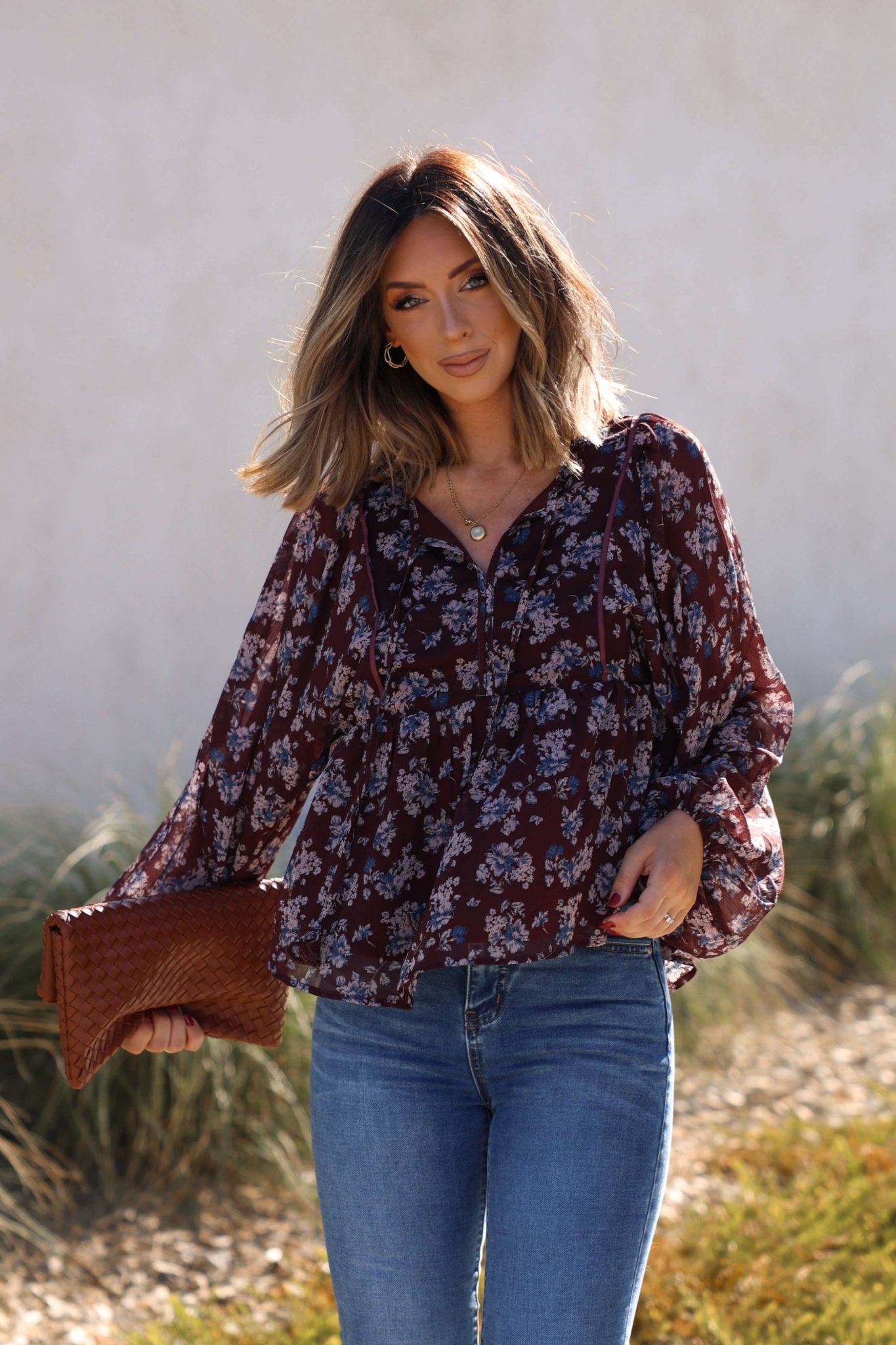 Woman outdoors in a Plum Blossom Babydoll Top and jeans, holding a brown woven clutch with grass in the background.