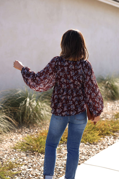 A woman wearing the Plum Blossom Babydoll Top and blue jeans stands outside with her back to the camera, holding a brown bag.