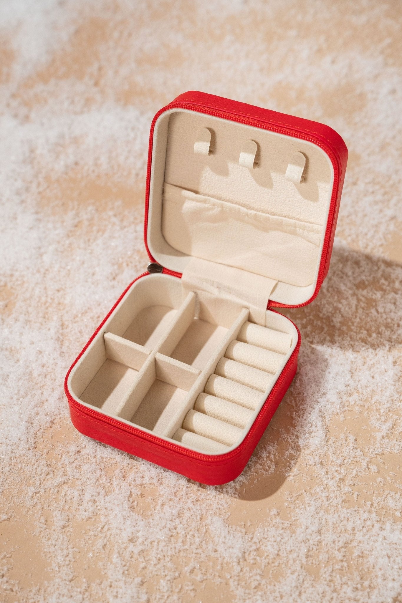 A Portable Red Leather Jewelry Box with compartments and ring holders sits open on a beige surface dusted with white powder.
