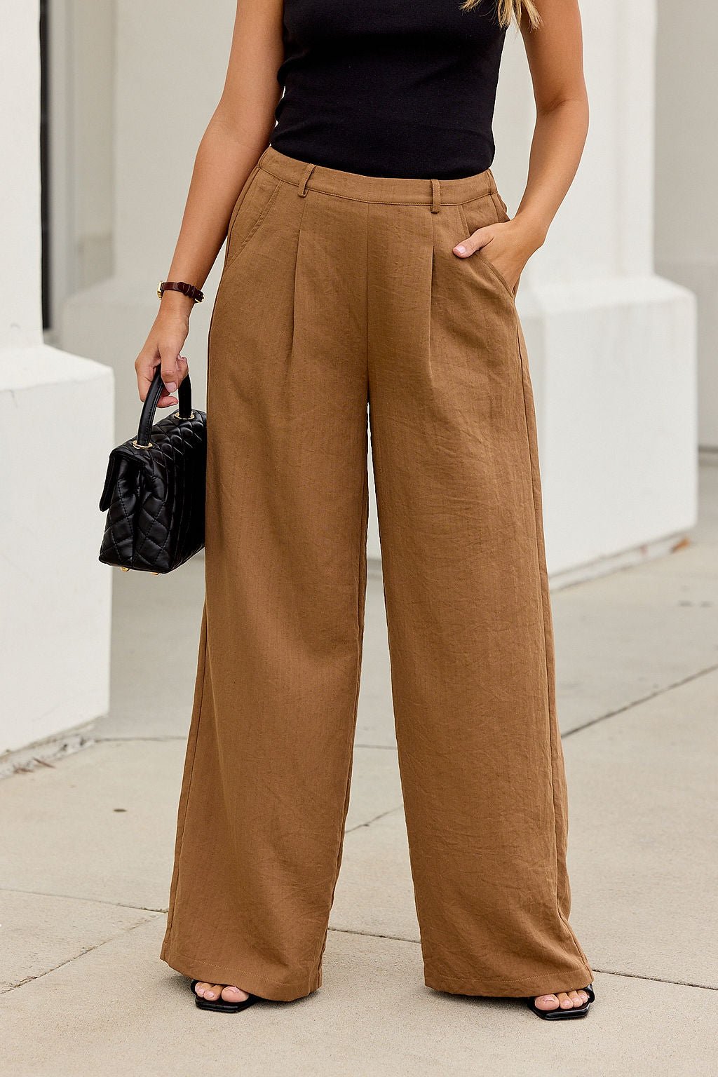 Woman in Power Pleated Wide Leg Pants - Camel, black sleeveless top, and sandals with a black handbag—a work-to-weekend staple.