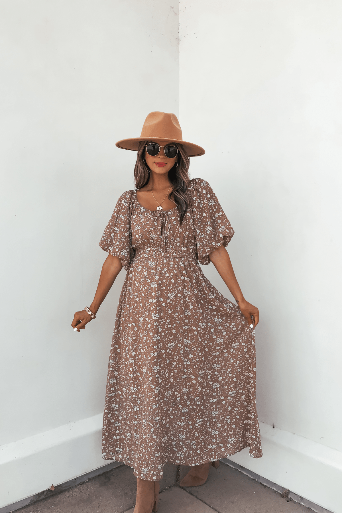 Woman in the Prairie Bloom Maxi Dress - Tan, tan hat, and sunglasses poses by a white wall, holding the dress sides.