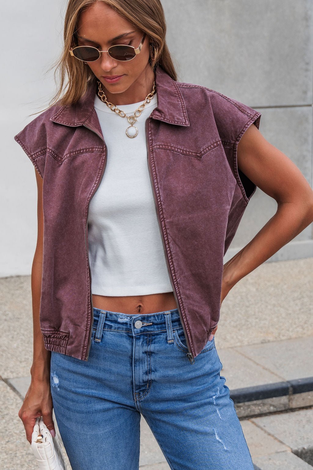Woman in sunglasses wears Presley Zip Up Collared Vest - Mauve, white crop top, blue jeans, and holds a white handbag outdoors.