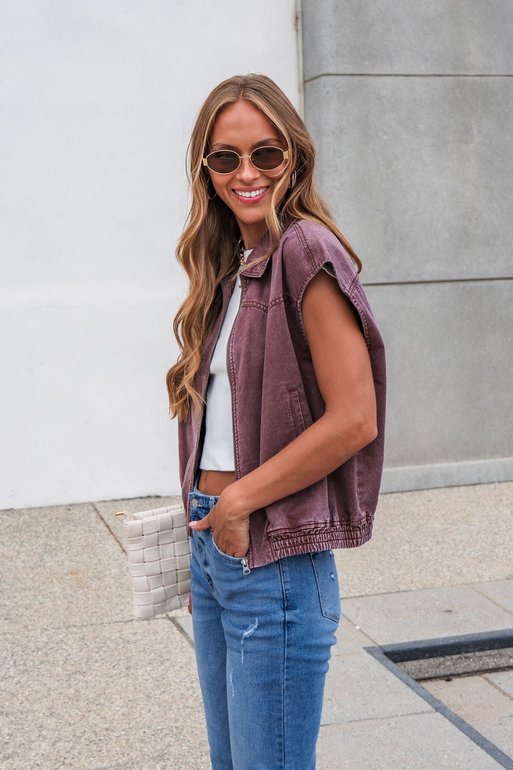 Woman with long hair in sunglasses smiles outdoors, wearing Presley Zip Up Collared Vest - Mauve, blue jeans, and holding a woven bag.