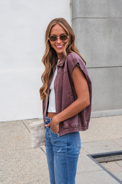 Woman with long hair in sunglasses smiles outdoors, wearing Presley Zip Up Collared Vest - Mauve, blue jeans, and holding a woven bag.