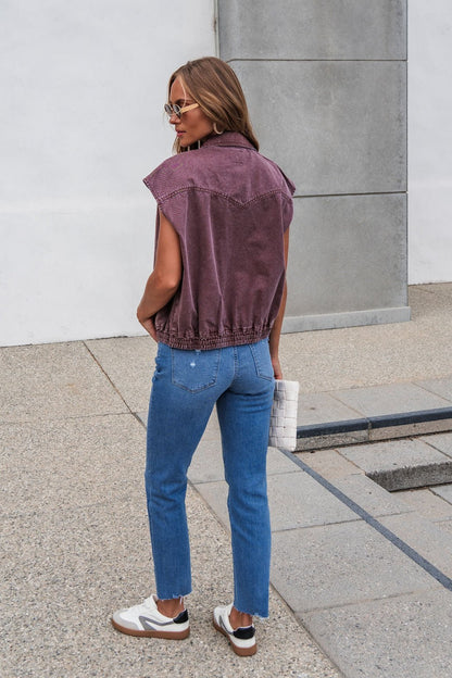 Woman in Presley Zip Up Collared Vest - Mauve, blue jeans, and white sneakers stands outdoors holding a white bag.