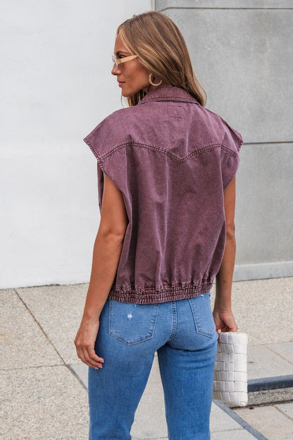 Woman in sunglasses wears the Presley Zip Up Collared Vest - Mauve, blue jeans, and holds a white woven clutch outdoors, facing away.