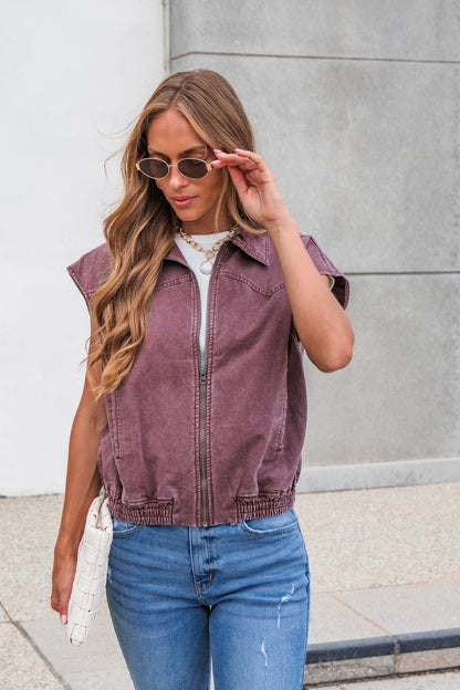 A woman in sunglasses and blue jeans wears the Presley Zip Up Collared Vest - Mauve, holding a white clutch outside.