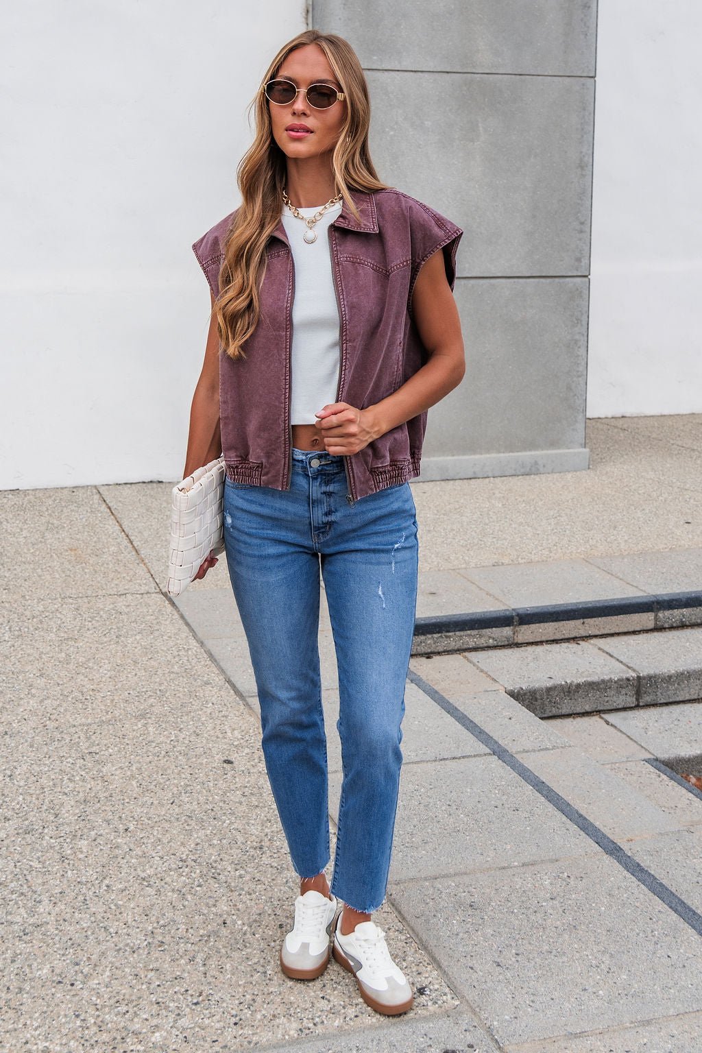 Woman in sunglasses wears the Presley Zip Up Collared Vest - Mauve with a white top, jeans, sneakers, and holds a clutch on a city sidewalk.