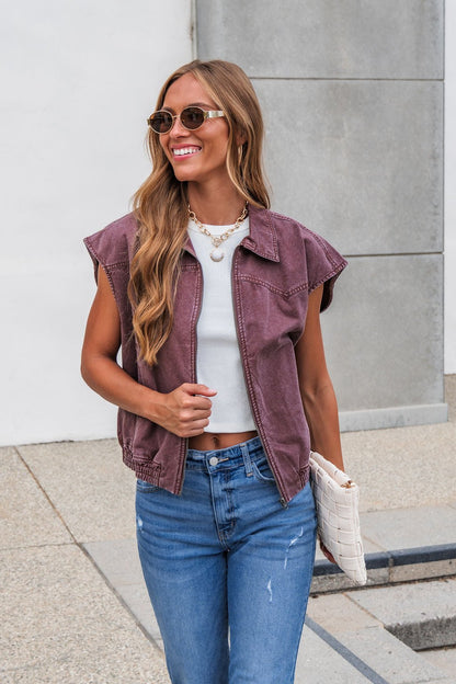 Woman with long hair in the Presley Zip Up Collared Vest - Mauve, white crop top, blue jeans, sunglasses, and clutch, smiling outdoors.