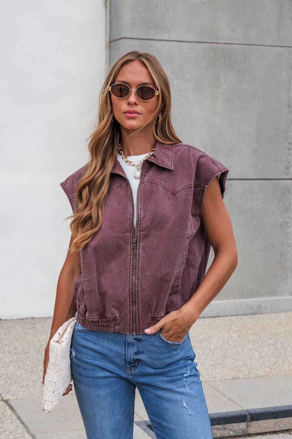 Woman in sunglasses wears the Presley Zip Up Collared Vest - Mauve with blue jeans and holds a white woven clutch outdoors.
