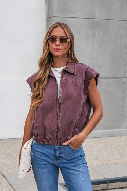Woman in sunglasses wears the Presley Zip Up Collared Vest - Mauve with blue jeans and holds a white woven clutch outdoors.