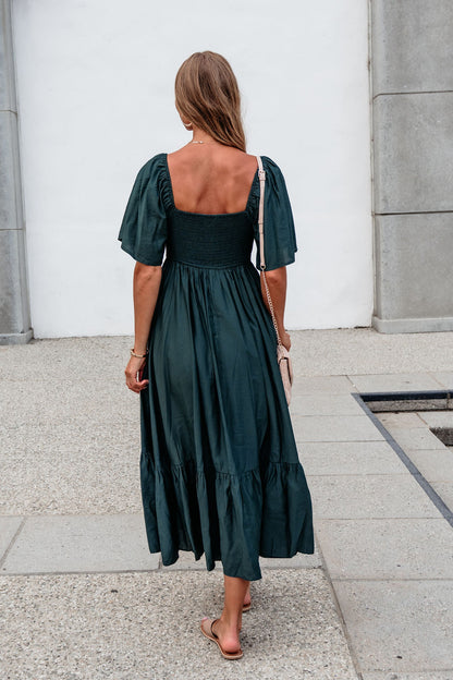 Woman in Rosalind Sweetheart Smocked Midi Dress — Green walks away on a paved path, carrying a light purse and wearing sandals.