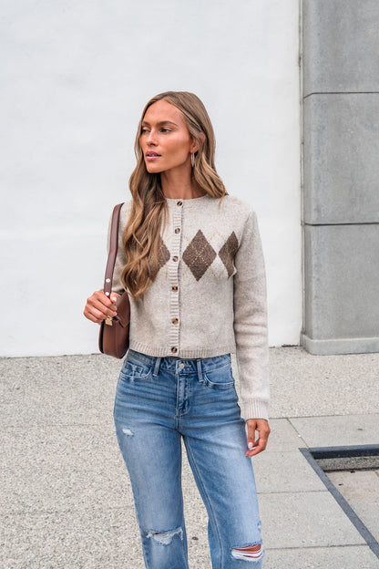 Woman with long hair wears The Wynn Diamond Cardigan - Taupe, ripped jeans, and a brown bag by a modern building.