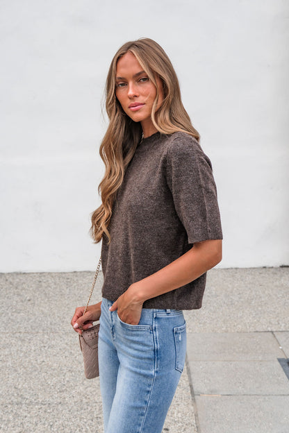Woman with long wavy hair wears the Tessa Mock Neck Sweater in brown and blue jeans, holding a small purse outdoors.
