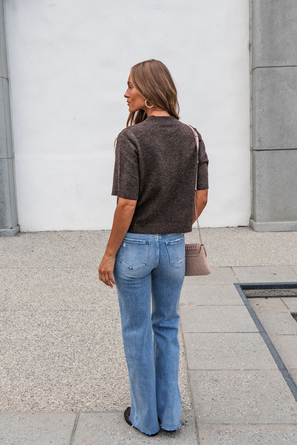 Woman with wavy hair in a brown top and Vervet Distressed Raw Hem Jeans - Light Wash stands on a paved area, facing away.