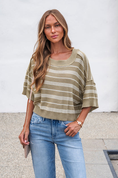 Woman in Bellamy Striped Linen Sweater - Olive and blue jeans stands outdoors holding a chain-strap purse against a white wall.