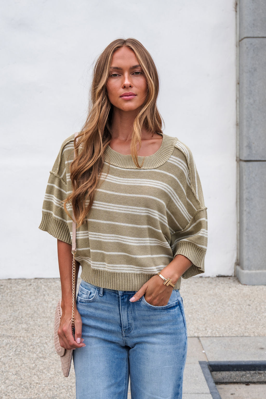 Woman with long hair wears the Bellamy Striped Linen Sweater - Olive and blue jeans, standing outdoors.