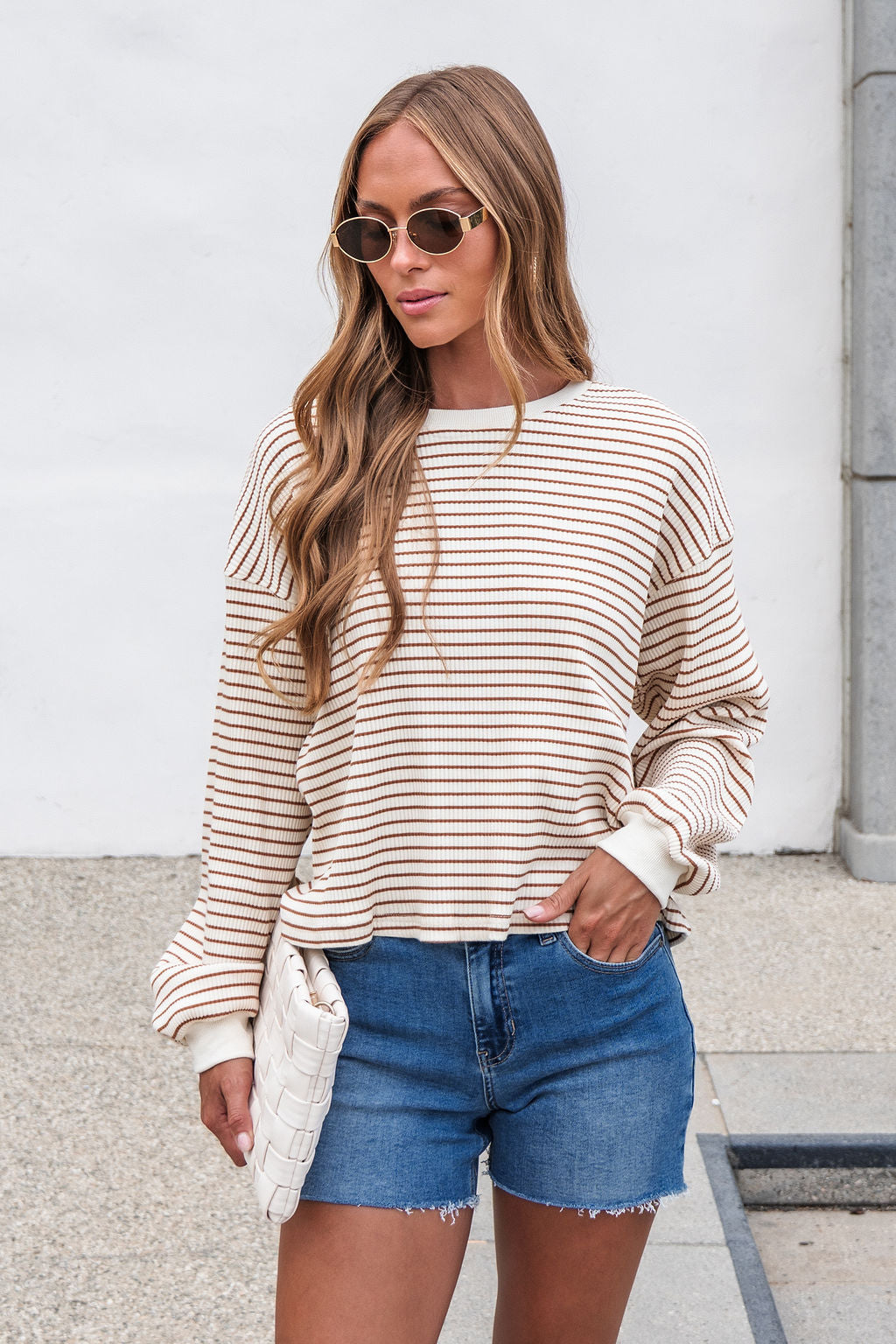 Woman in sunglasses wears the Sutton Striped Pullover - Cream and denim shorts, holding a white clutch outdoors.