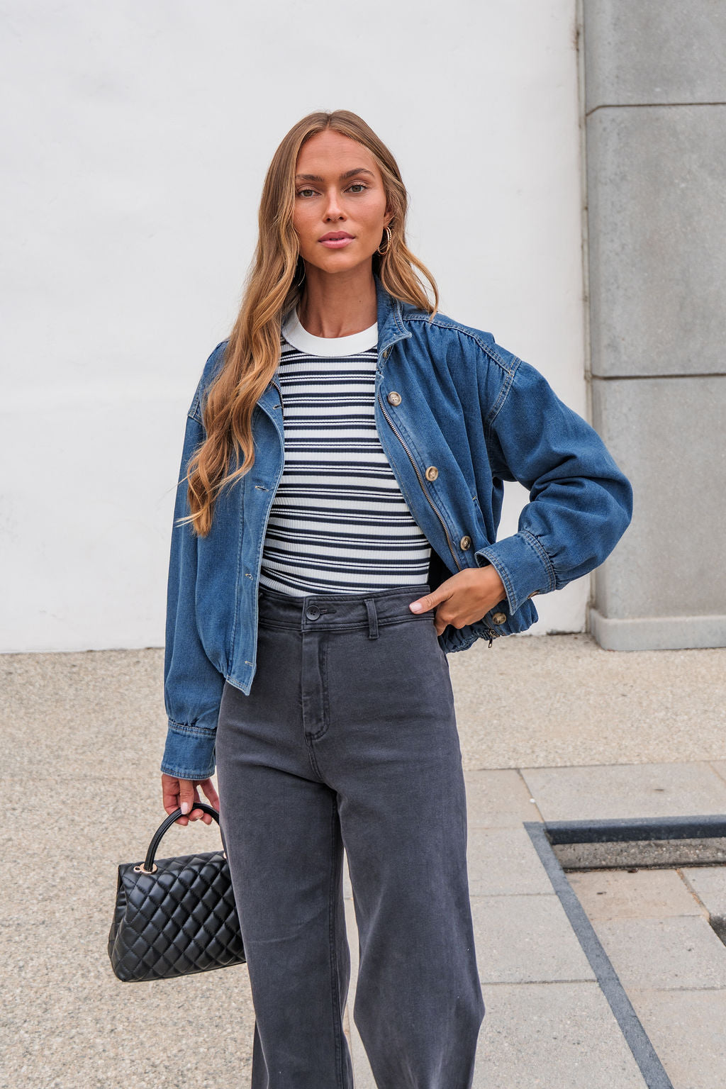A woman in The Dakota Denim Jacket, striped shirt, and black jeans stands outdoors holding a small black quilted handbag.