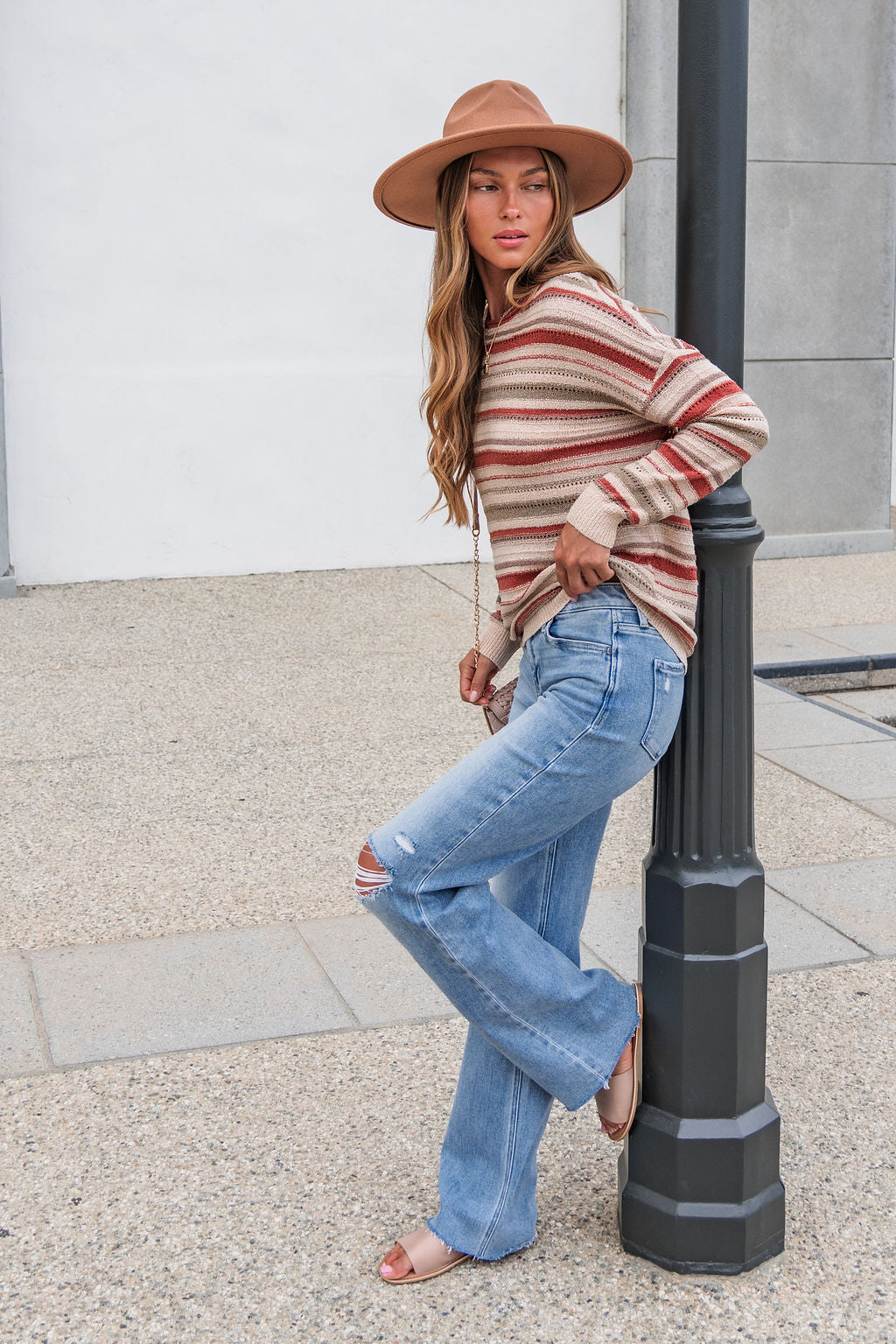Woman wearing The Reeves Striped Sweater - Red, blue jeans, and a hat leans against a lamp post on the sidewalk, gazing to the side.
