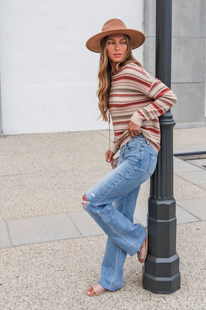 Woman wearing The Reeves Striped Sweater - Red, blue jeans, and a hat leans against a lamp post on the sidewalk, gazing to the side.