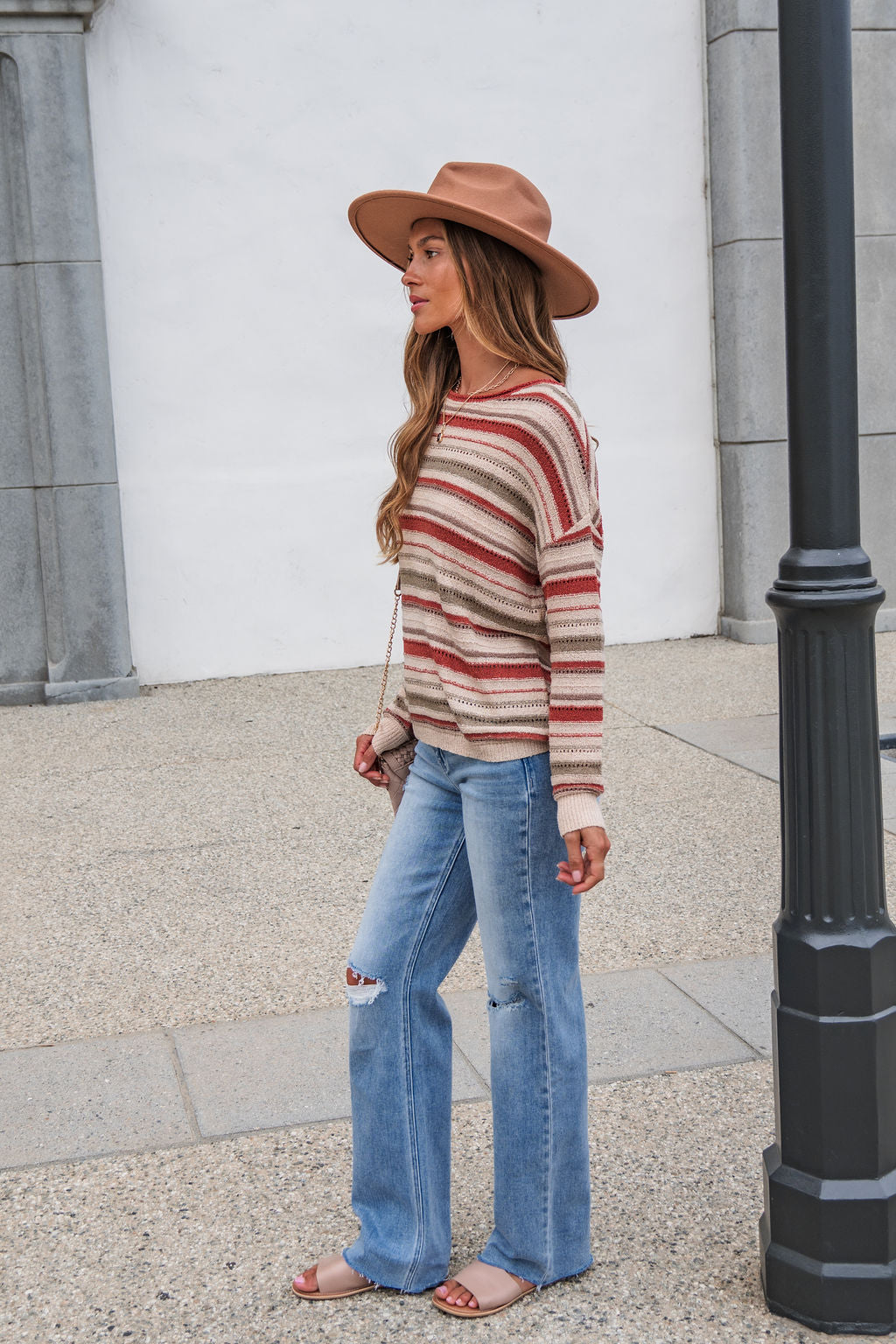 Woman in The Reeves Striped Sweater - Red, jeans, hat, and sandals stands by a lamp post on a gray sidewalk with white walls.