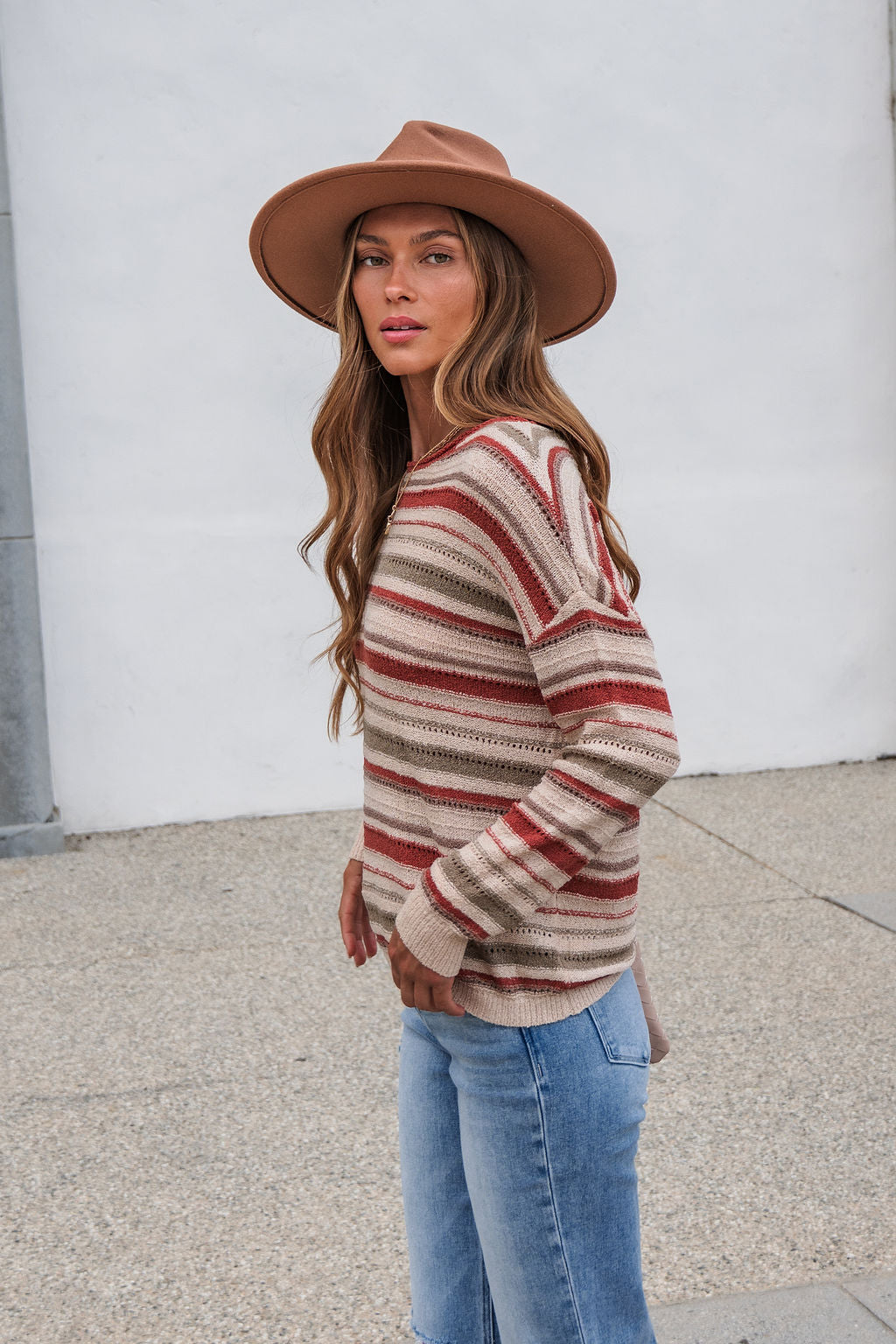 Woman in The Reeves Striped Sweater - Red, jeans, and a brown hat stands outdoors against a neutral wall.