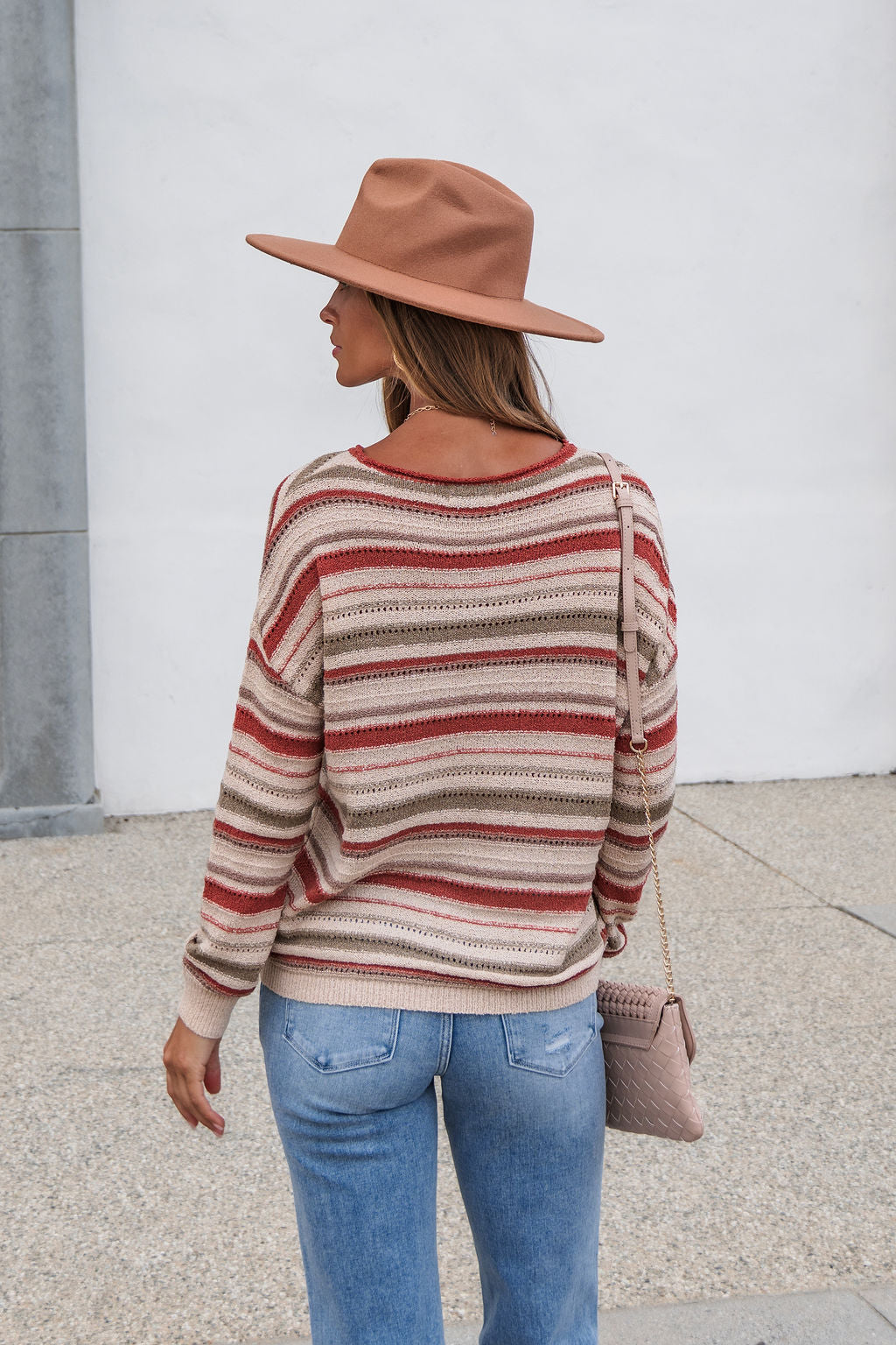 Someone in The Reeves Striped Sweater - Red, jeans, brown hat, and crossbody purse stands outside facing away from the camera.