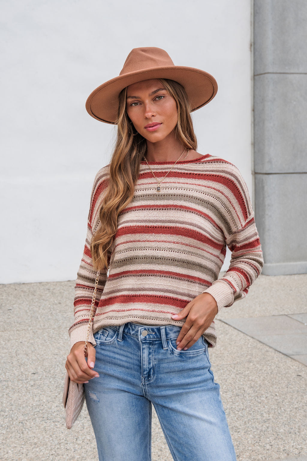 Woman wears The Reeves Striped Sweater - Red, a tan hat, and blue jeans while standing outdoors against a light wall and concrete.