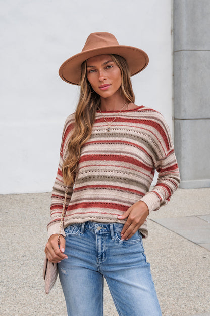 Woman wears The Reeves Striped Sweater - Red, a tan hat, and blue jeans while standing outdoors against a light wall and concrete.