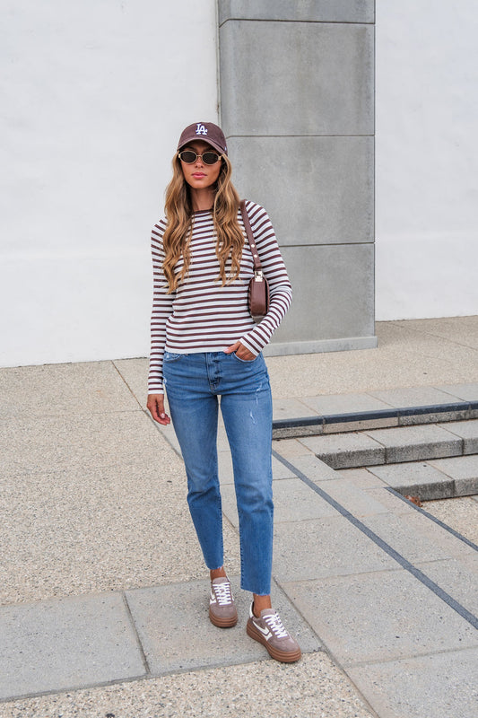 Woman in The Josephine Striped Top - Brown, blue jeans, brown sneakers, and hat stands on a concrete path holding a small bag.