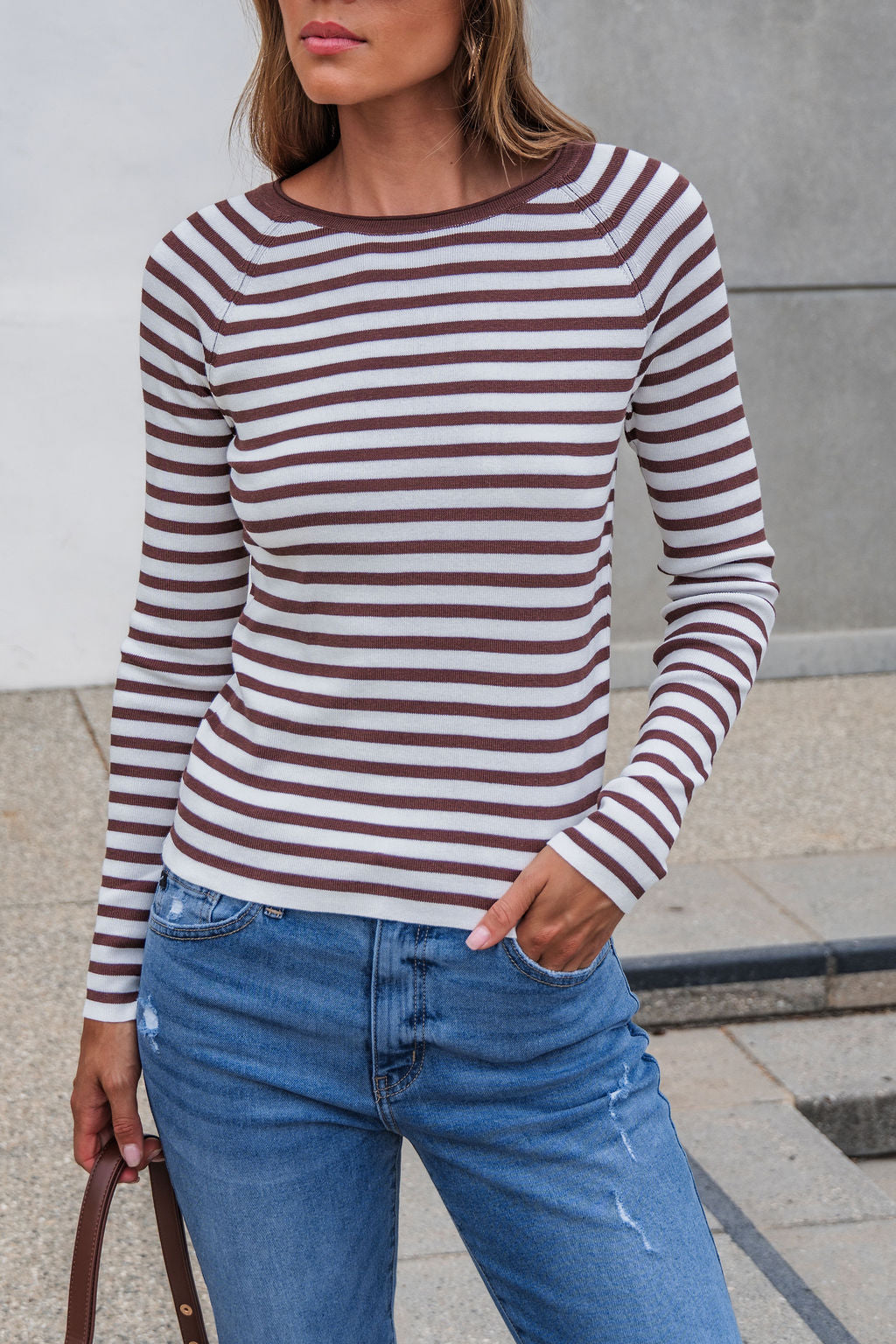 Woman wears The Josephine Striped Top - Brown with ribbed detail and blue jeans, standing outdoors with one hand in her pocket.