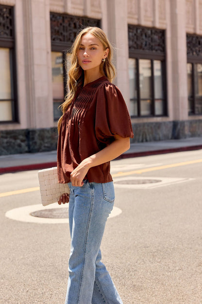 A woman in a brown Puff Sleeve Tie Front Top and blue jeans stands on a city street, holding a white clutch, looking at the camera.