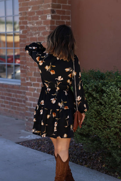 A woman wears the Pumpkin Patch Black Floral Print Mini Dress and brown boots, standing on a sidewalk with her hand in her hair.