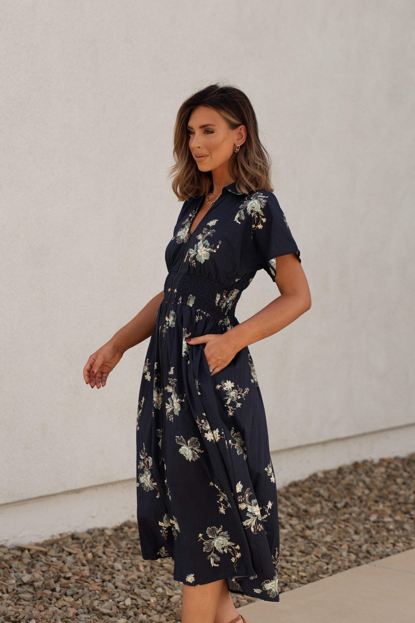 Woman in a Pumpkin Spice Black Floral Zip Midi Dress stands outdoors on a stone path by a white wall, looking off to the side.