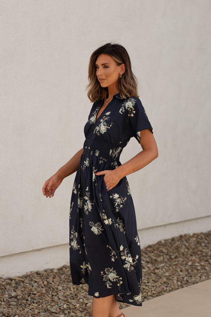 Woman in a Pumpkin Spice Black Floral Zip Midi Dress stands outdoors on a stone path by a white wall, looking off to the side.