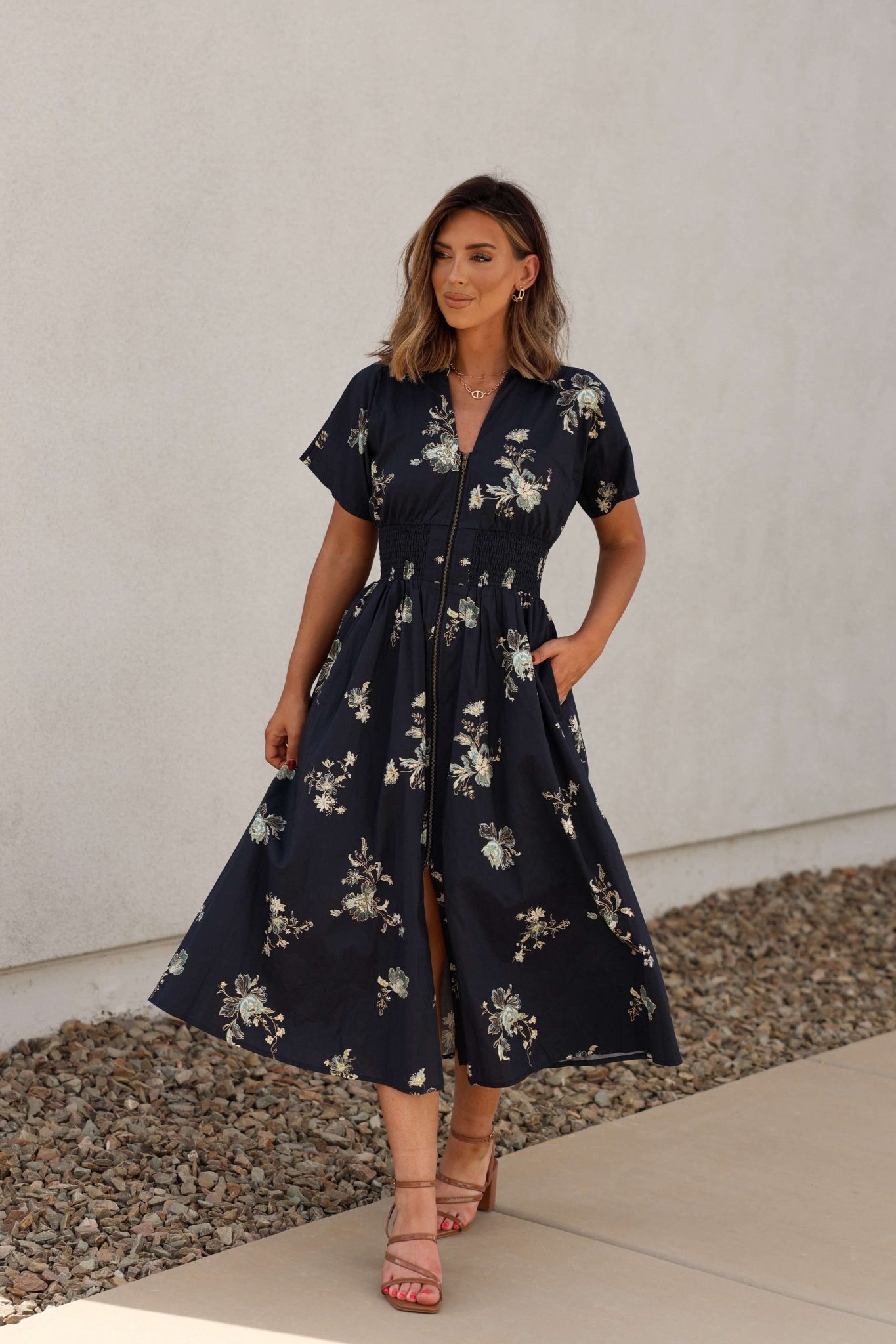 Woman in a Pumpkin Spice Black Floral Zip Midi Dress stands on a sidewalk near a white wall and rocky ground.